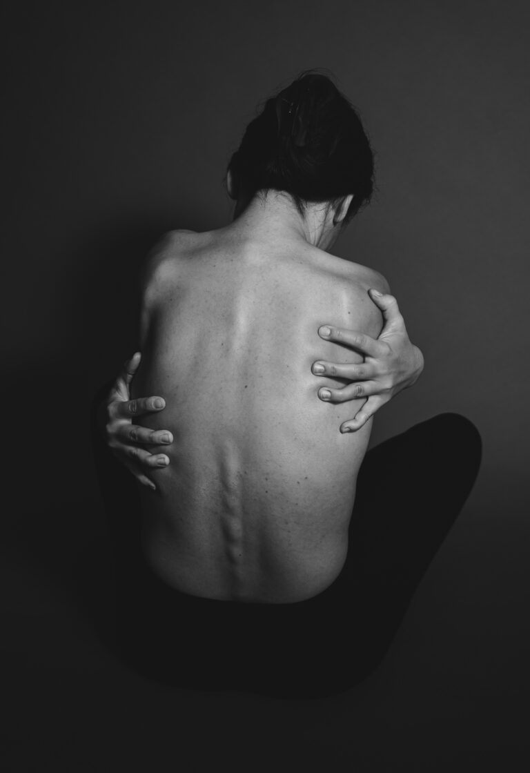 A black-and-white photograph of Petra sitting with her back turned, holding herself with both hands.