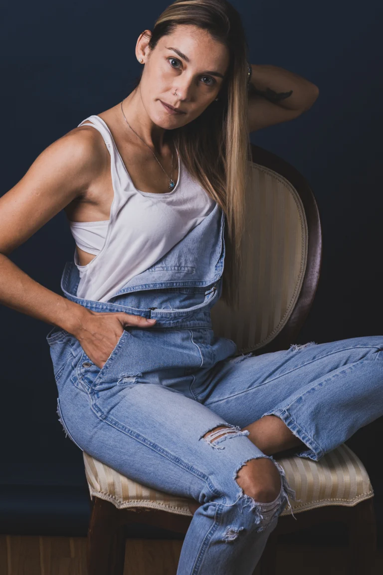 A woman with long blonde hair, wearing a white tank top and ripped denim overalls, sits on an upholstered chair against a dark background.