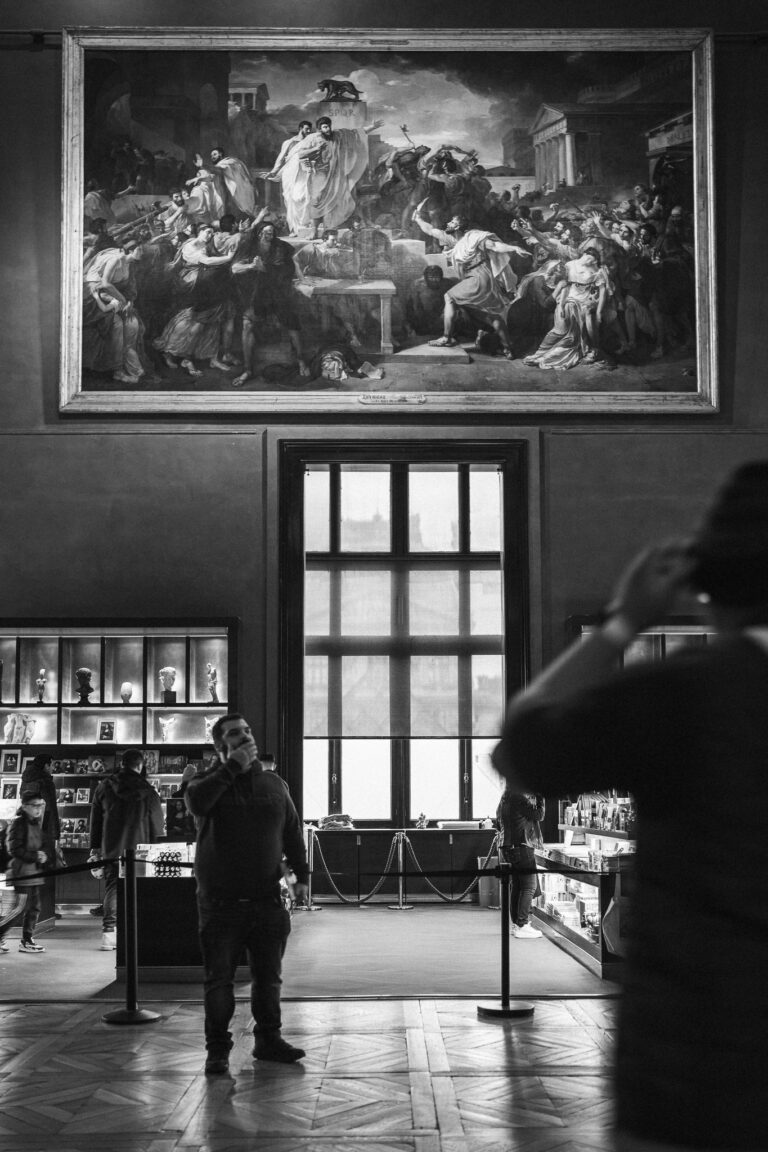Ein Mann steht in einem Museumssaal vor einem großen Gemälde an der Wand über einem Fenster, während eine andere Person im Vordergrund silhouettiert ist.