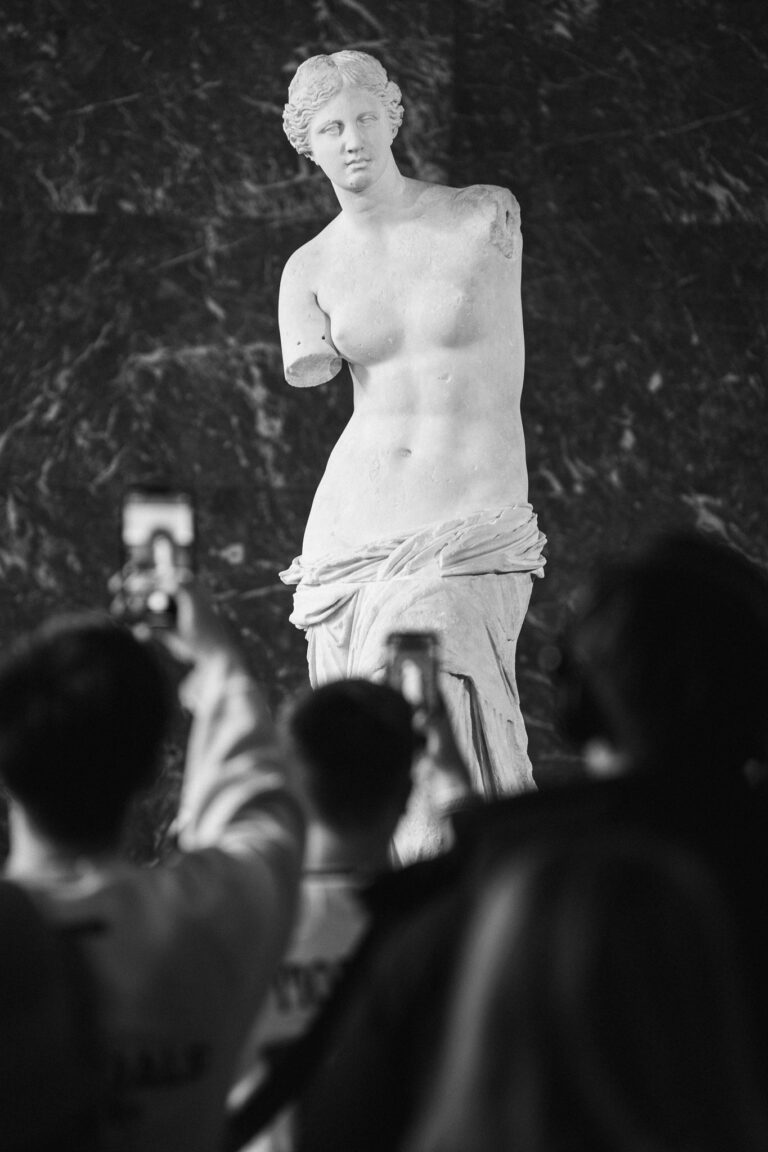 Monochrome photograph of museum visitors photographing the Venus de Milo statue with their smartphones.