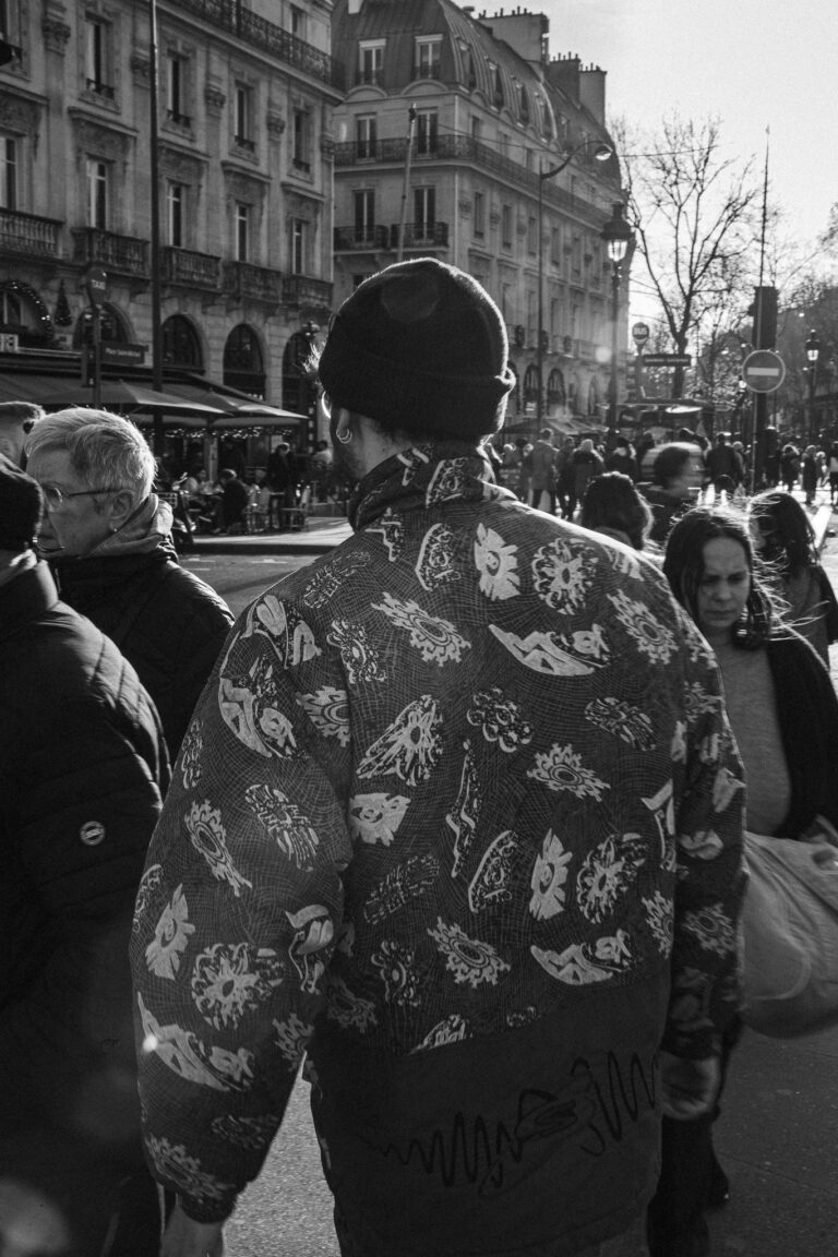 Eine Schwarz-Weiß-Straßenszene zeigt einen Mann mit gemusterter Jacke und Mütze, der durch eine Menschenmenge in Paris geht.