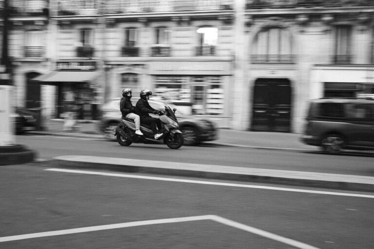 Eine schwarz-weiße Mitzieher-Aufnahme in Paris, die einen Mann und eine Frau mit Helmen auf einem Roller auf einer Straße zeigt.