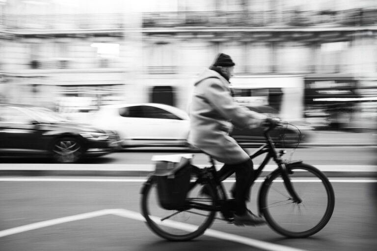 A man rides a bicycle past blurred urban buildings and cars in a motion-blurred black and white street photograph.
