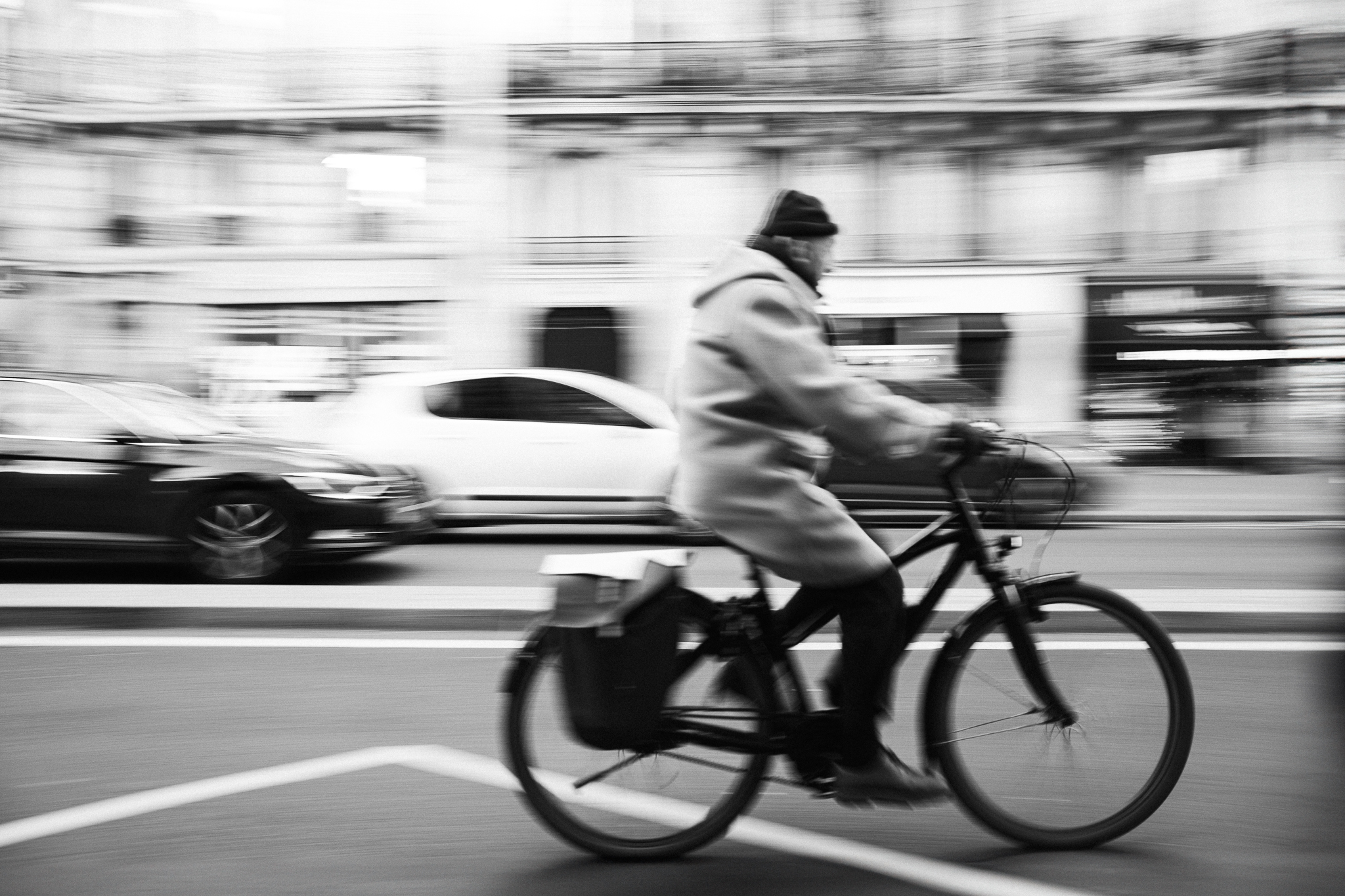 Ein Mann fährt in einer verwischten Schwarz-Weiße-Straßenszene auf einem Fahrrad an verschwommenen Gebäuden und Autos vorbei.