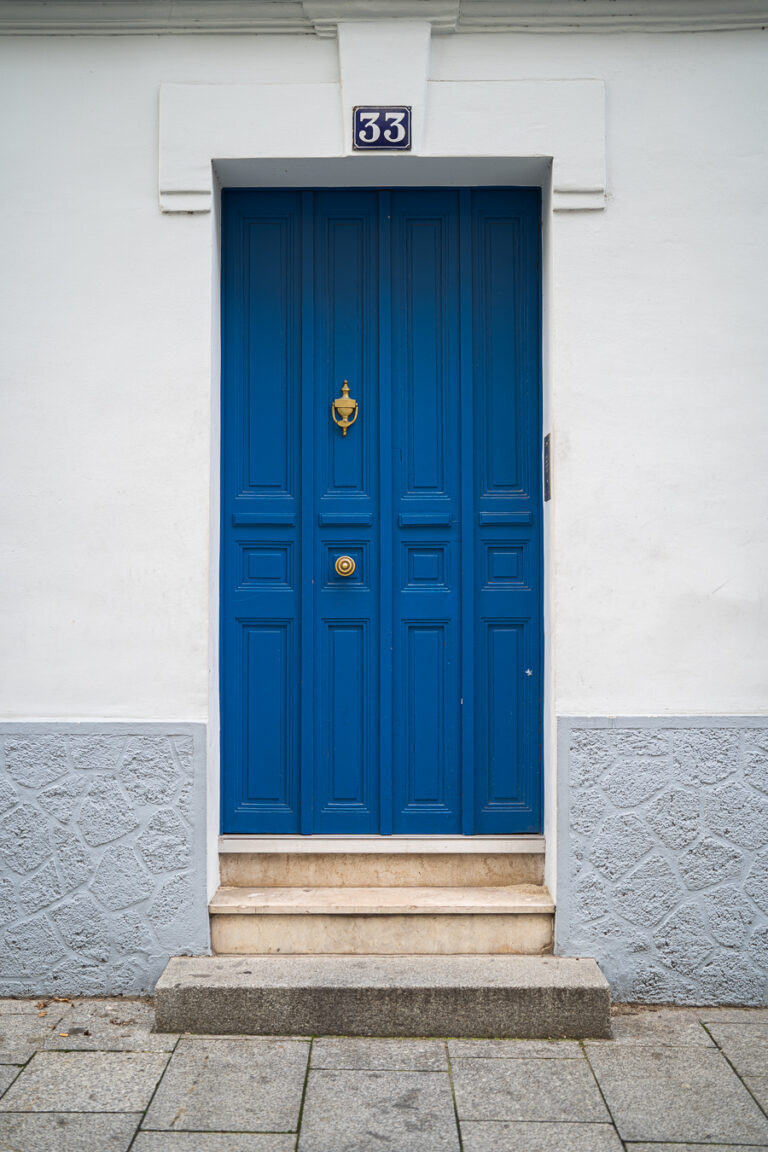 Eine leuchtend blaue Kassettentür mit goldenen Beschlägen und der Nummer 33 darüber, eingelassen in eine weiße Wand mit grauem Steinsockel.