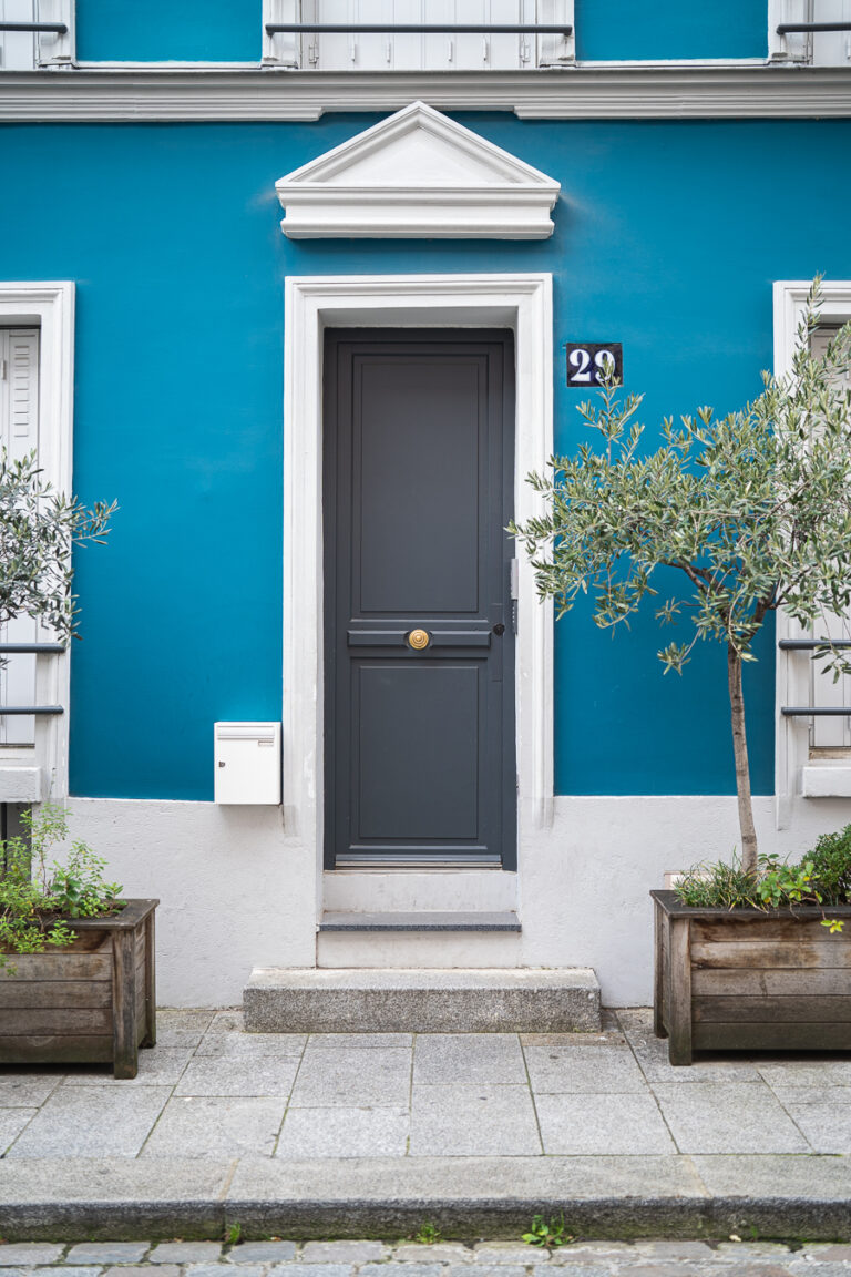 A dark grey panelled door with a round brass knob, centered in a bright blue building facade with a number 29 plaque and flanked by potted olive trees.