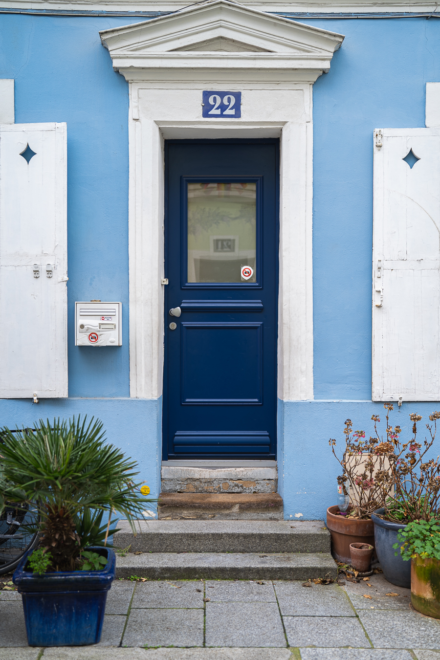 Eine dunkelblaue Haustür in einer hellblauen Fassade, flankiert von weißen Fensterläden, Topfpflanzen und Steinstufen.