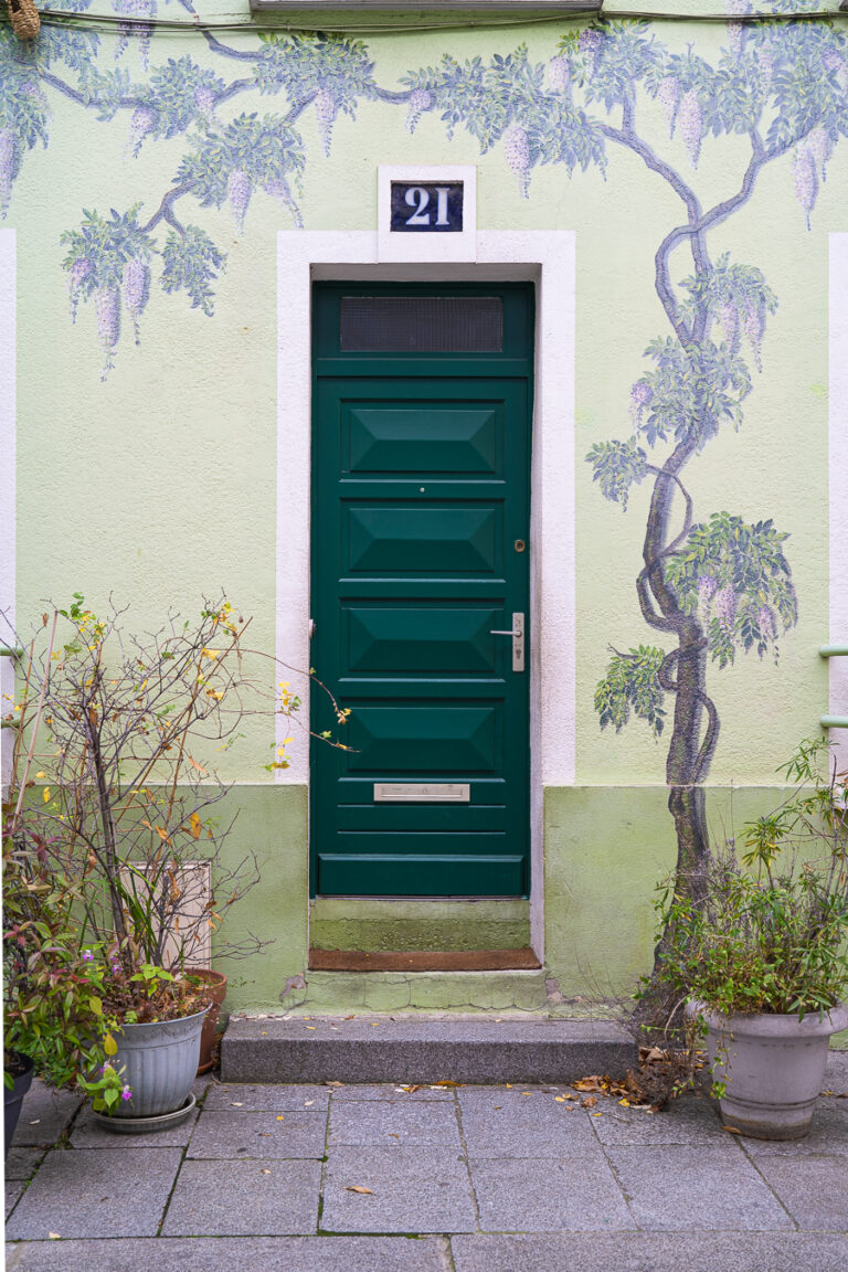 Eine dunkelgruene Kassettentuer mit der Hausnummer 21 befindet sich unter einem gemalten Wisterien-Wandgemalde an einer hellgruenen Fassade.
