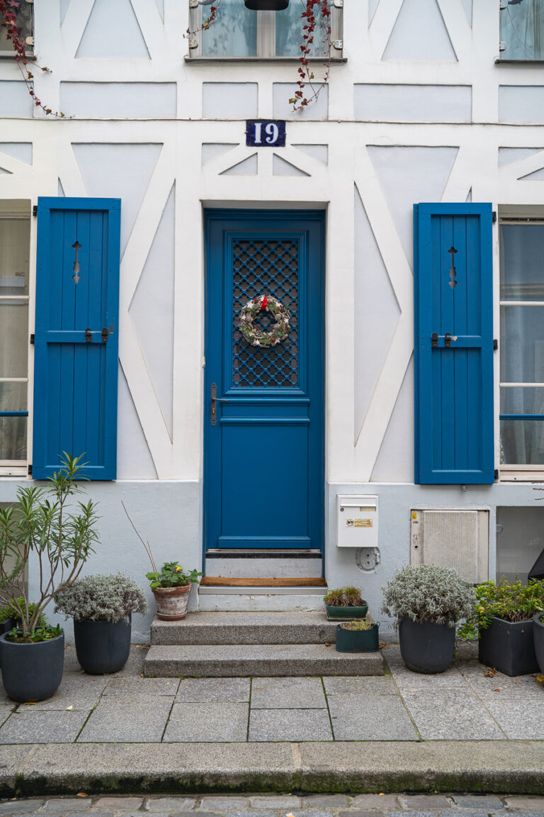 Eine hellblaue Holzeingangstür mit einem kleinen Weihnachtskranz unter der Hausnummer 19, flankiert von passenden blauen Fensterläden und Topfpflanzen auf den Steinstufen.