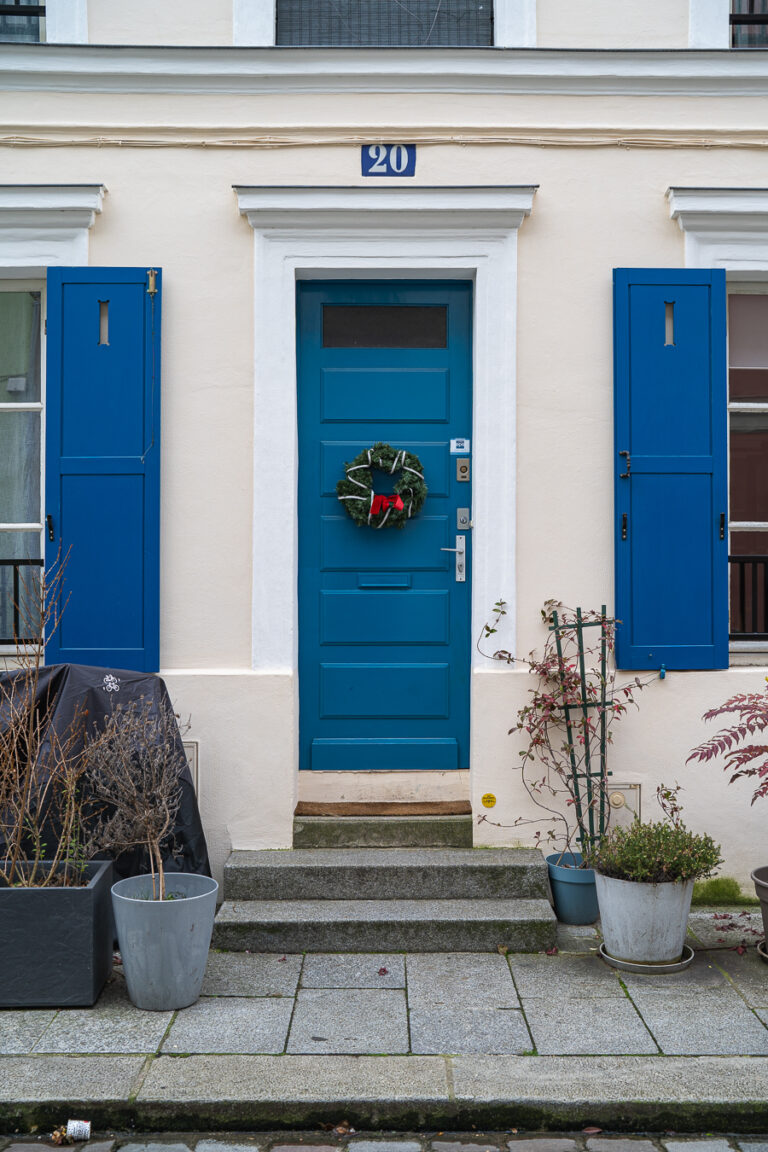 Eine hellblaue Haustür mit einem weihnachtlichen Kranz, umgeben von blauen Fensterläden und Topfpflanzen, unterhalb der Hausnummer 20.
