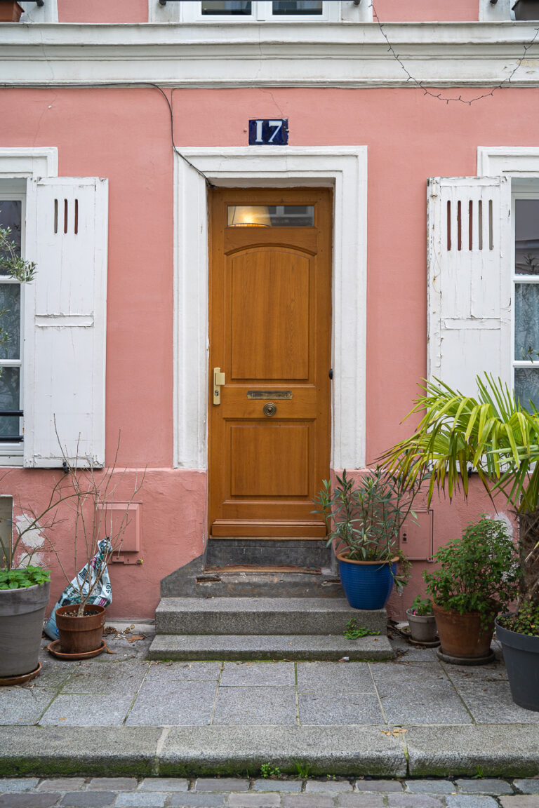 A wooden front door with a number 17 sign above it, set in a pink building facade with white shutters and potted plants on the stone pavement.