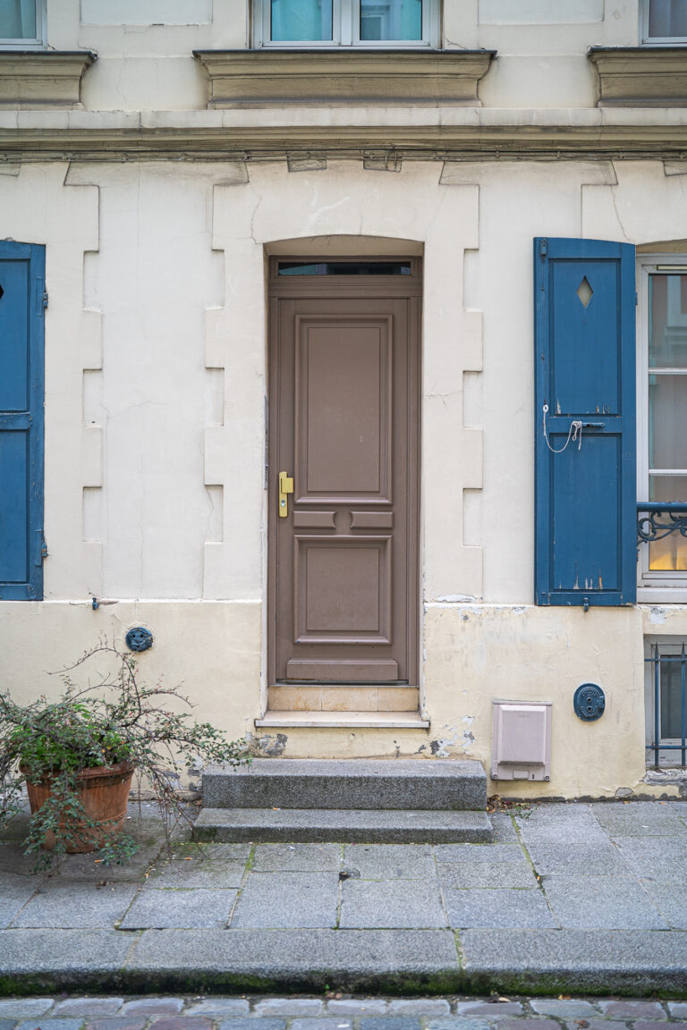 Eine braune Kassettentür in einer hellen Fassade mit blauen Fensterläden an den Nachbarfenstern und zwei Steinstufen davor.