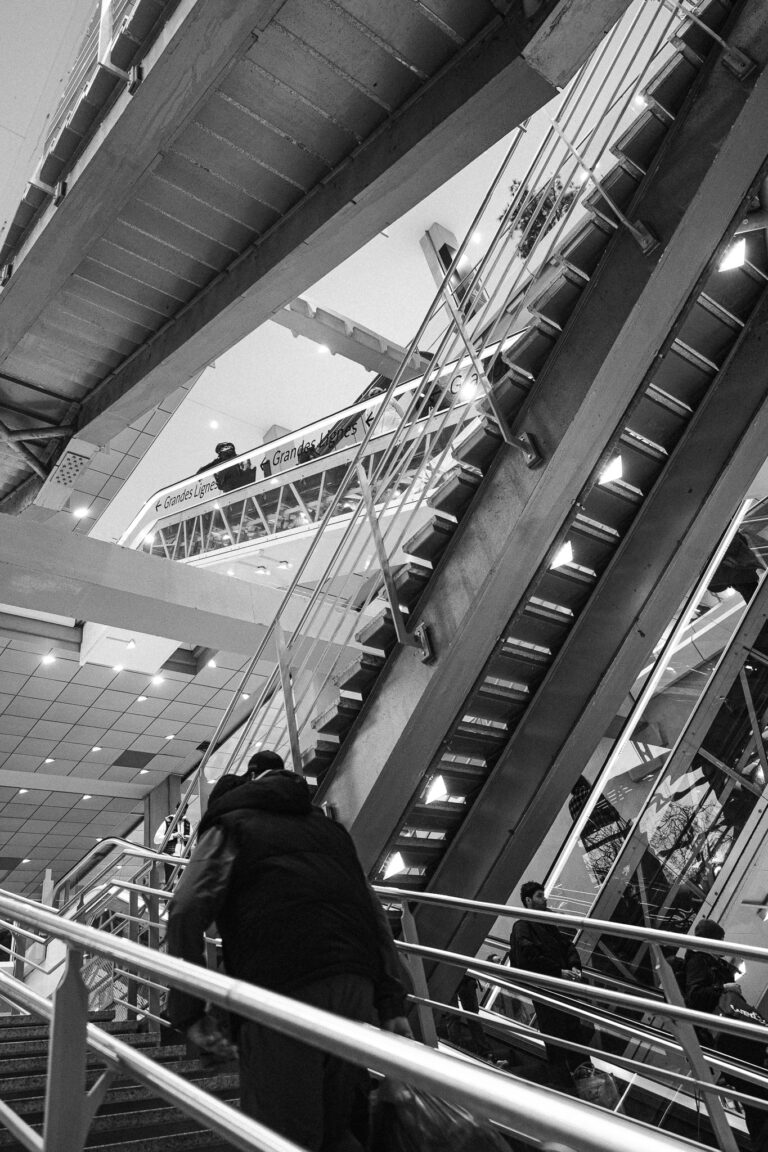 Low-angle black and white shot of a man walking up a modern train station staircase with overlapping ramps and lit steps.