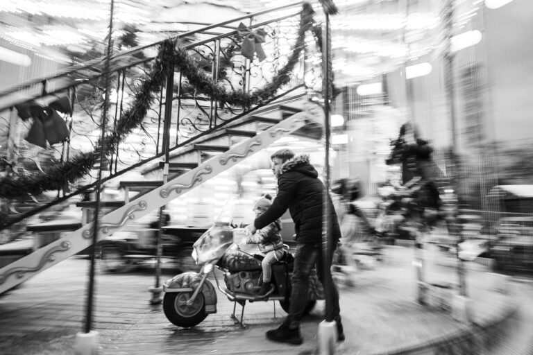 A black-and-white motion-blurred shot of a man standing beside a child seated on a carousel motorcycle.