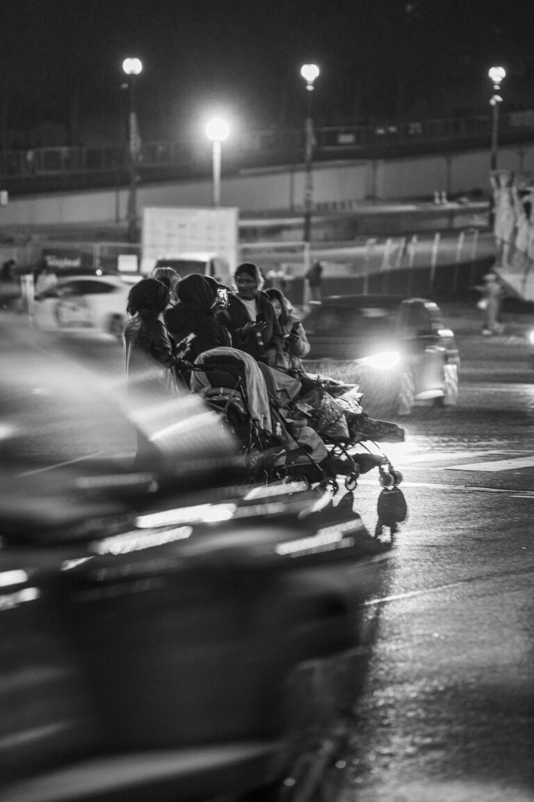 Eine nächtliche Schwarz-Weiß-Straßenszene in Paris zeigt Menschen mit einem Kinderwagen beim Überqueren einer nassen Straße mit verschwommenem Verkehr.