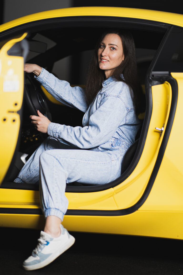 Petra sits in the driver seat of a yellow car, holding the steering wheel and looking toward the camera.