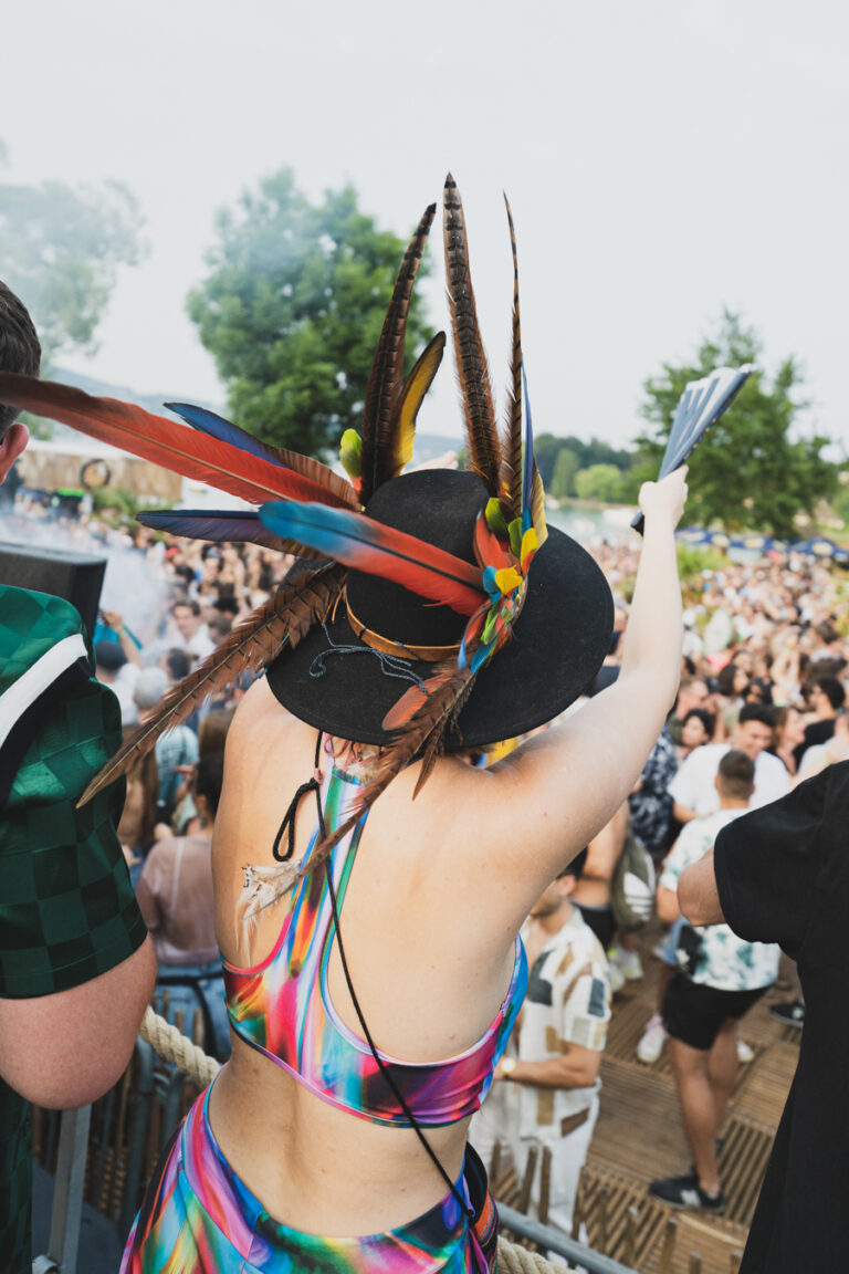 Eine Frau mit einem schwarz, mit bunten Federn geschmückten Hut steht inmitten einer verschwommenen Festivalmenge und hält einen Fächer in die Höhe.