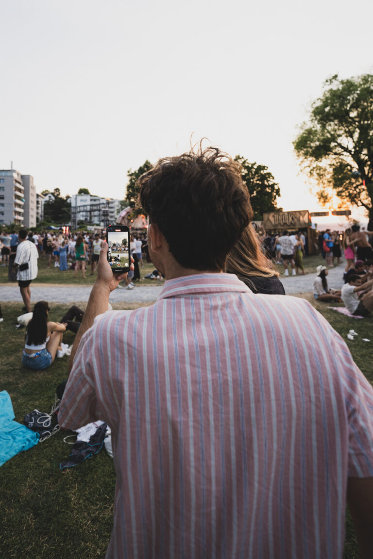 Ein Mann im gestreiften Hemd hält ein Smartphone hoch, um eine Menschenmenge in einem Park zu filmen.