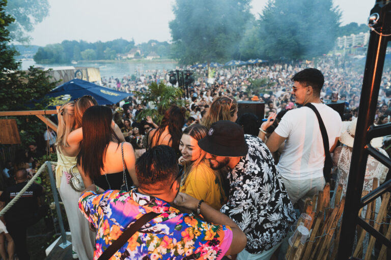 Eine große Menge von Männern und Frauen versammelt sich auf einem Freiluftfestival an einem See, während sich im Vordergrund Menschen unterhalten.