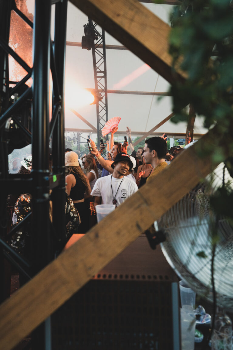 Eine Menschenmenge unter einem Pavillon auf einem Festivalgelände, eingerahmt von Holzbalken und Metallgerüsten, mit einem Mann im Vordergrund in weißem Hemd und dunklem Anglerhut.
