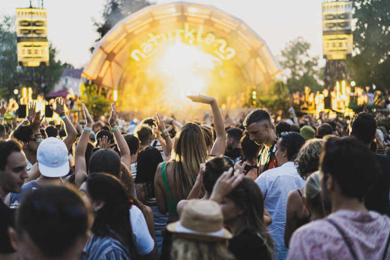 Eine grosse Menschenmenge mit erhobenen Händen versammelt sich bei Sonnenuntergang vor einer beleuchteten Open-Air-Festivalbühne.