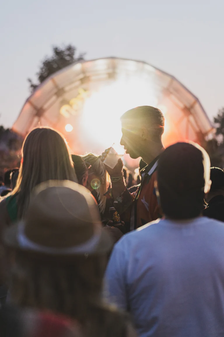 A crowd of people stands outdoors facing a brightly lit stage during sunset, with a man drinking from a cup in the center.