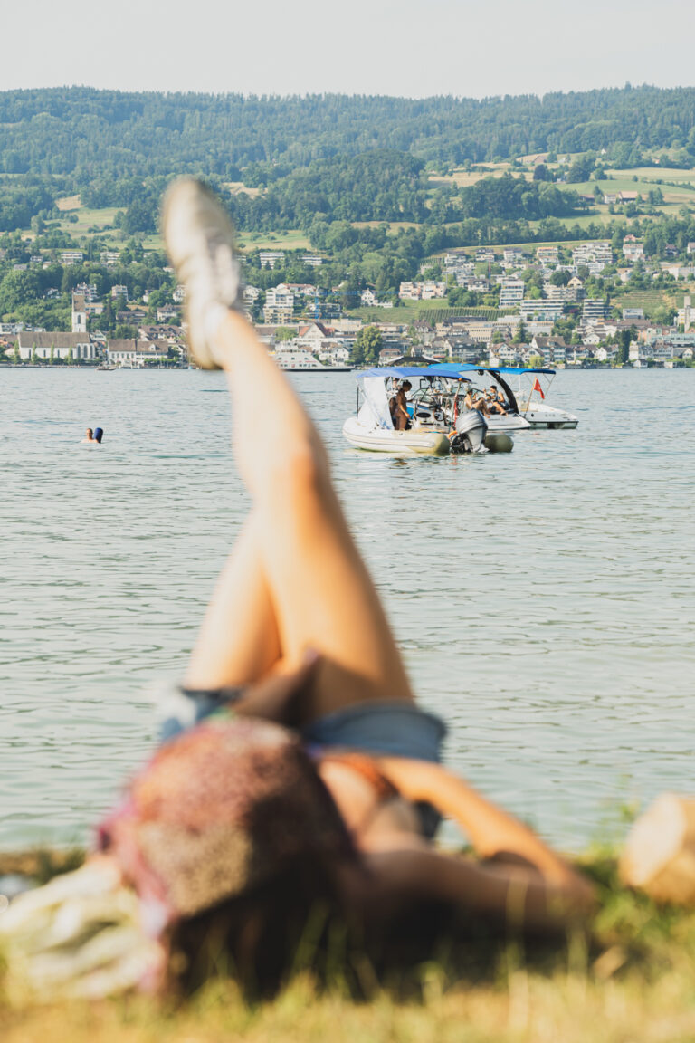 Eine Frau liegt im Gras an einem See, wobei ihre Beine im Vordergrund vor einer Kulisse aus Wasser, Booten und einer fernen Stadt nach oben gehalten werden.