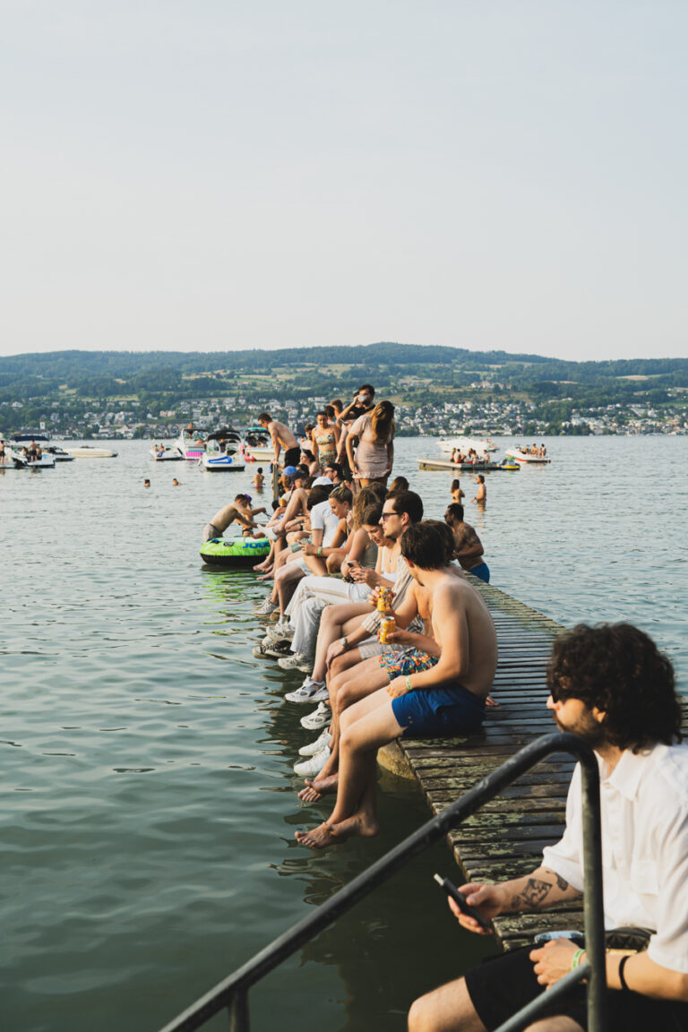 Männer und Frauen sitzen auf einem Holzsteg, der in einen ruhigen See ragt, mit fernen Hügeln und Booten bei natürlichem Tageslicht.