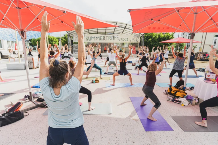 A large group of people practicing yoga outdoors on mats under red parasols, with a banner reading SUMMERTRAFF VADOZ in the background.