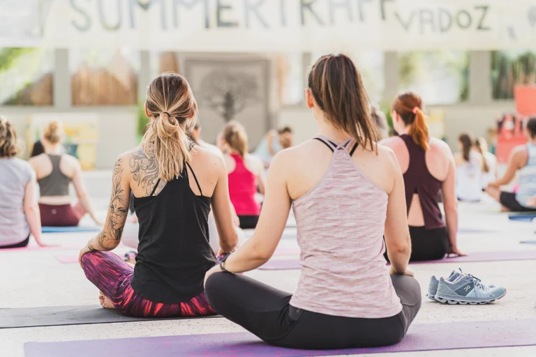 Several women sit cross-legged on yoga mats in an outdoor yoga class, viewed from behind.