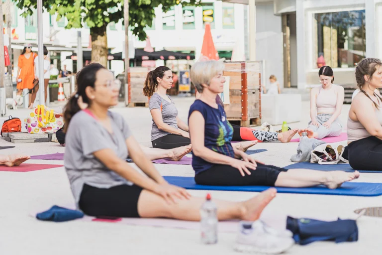 Several women sit on yoga mats outdoors on a paved public square.