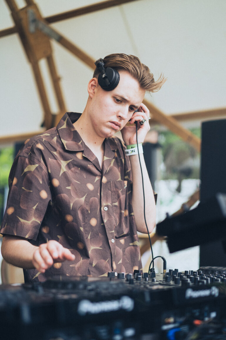 Ein Mann in einem gemusterten Kurzarmhemd steht hinter einem DJ-Mixer und hält sich bei Tageslicht Kopfhörer an ein Ohr.