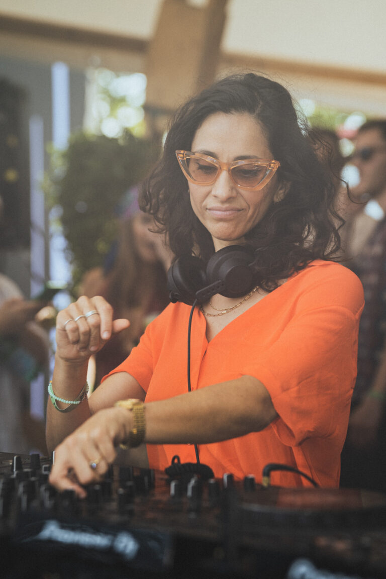 Eine Frau mit orangefarbener Sonnenbrille und einem orangefarbenen Oberteil bedient im Freien bei natürlichem Licht ein DJ-Mischpult mit Kopfhörern um den Hals.