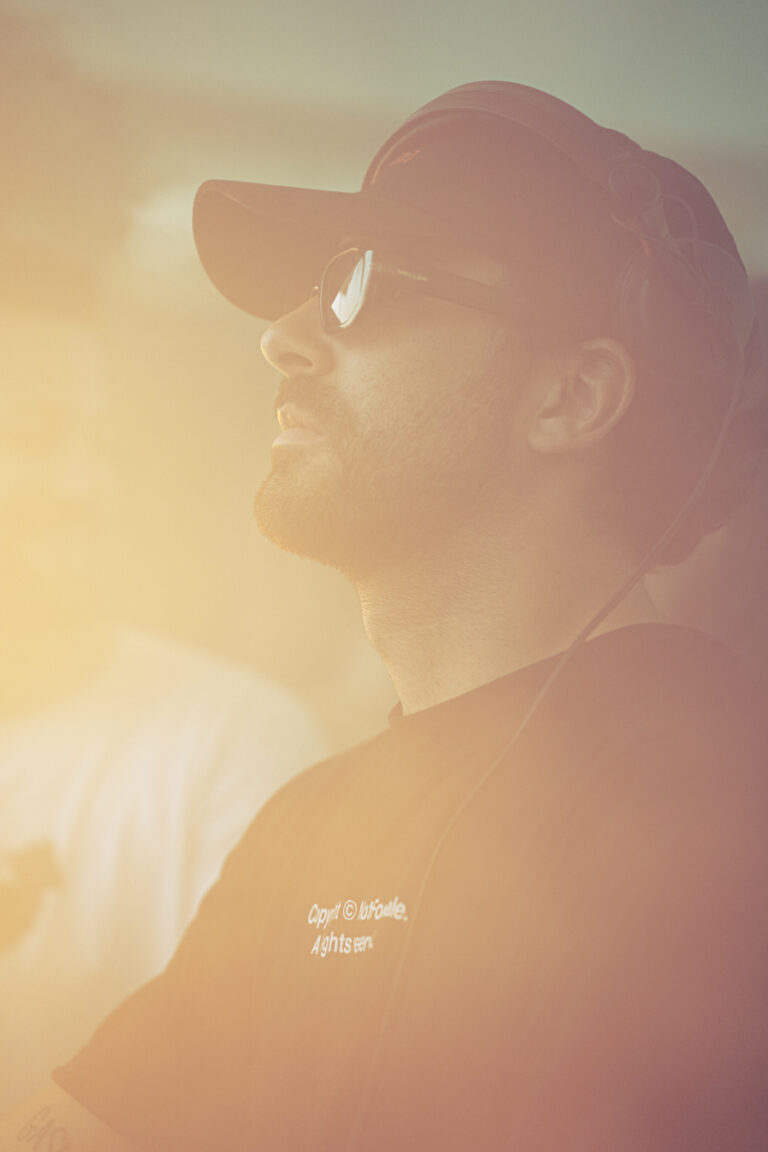 Profile view of a bearded man wearing a cap and sunglasses, captured in hazy, sun-drenched warm lighting.