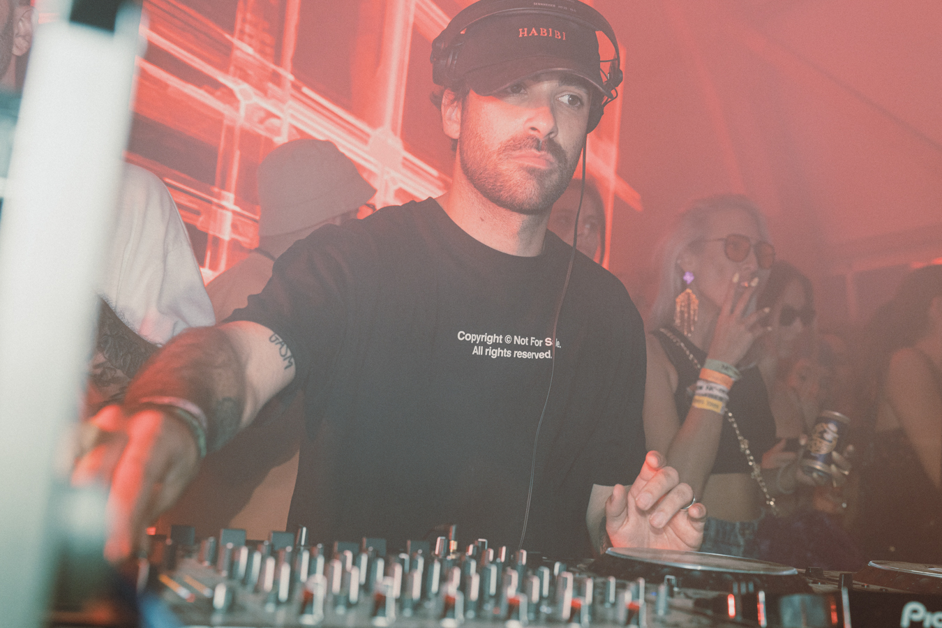 A man wearing a black cap and t-shirt stands behind a mixing console under red lighting, with a crowd visible in the background.