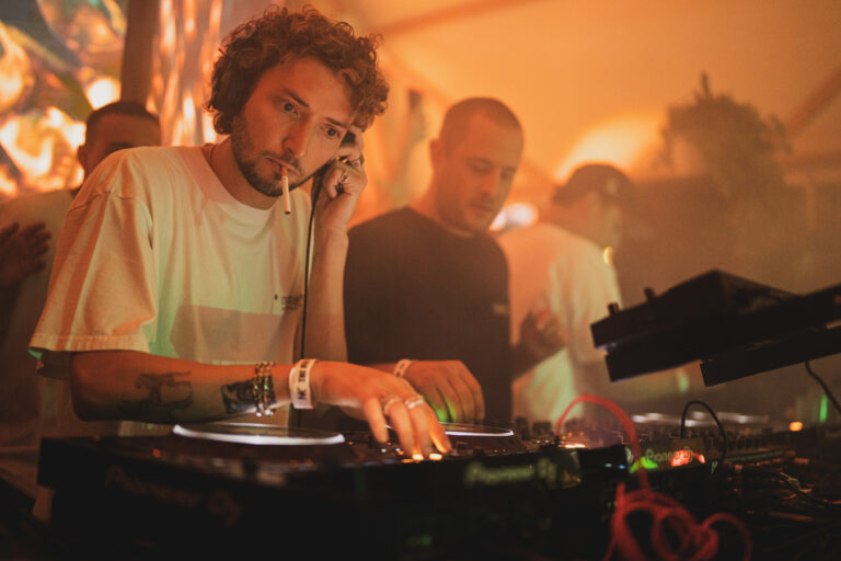 Ein lockiger Mann mit einer Zigarette im Mund steht unter warmem orangem Licht an einem DJ-Mixer, während andere Männer im Hintergrund zu sehen sind.