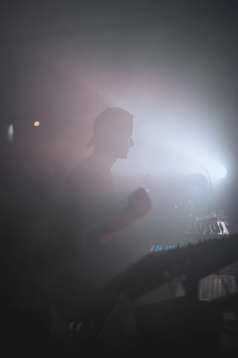 A man wearing a cap operates a mixing console in a hazy room illuminated by bright stage light.