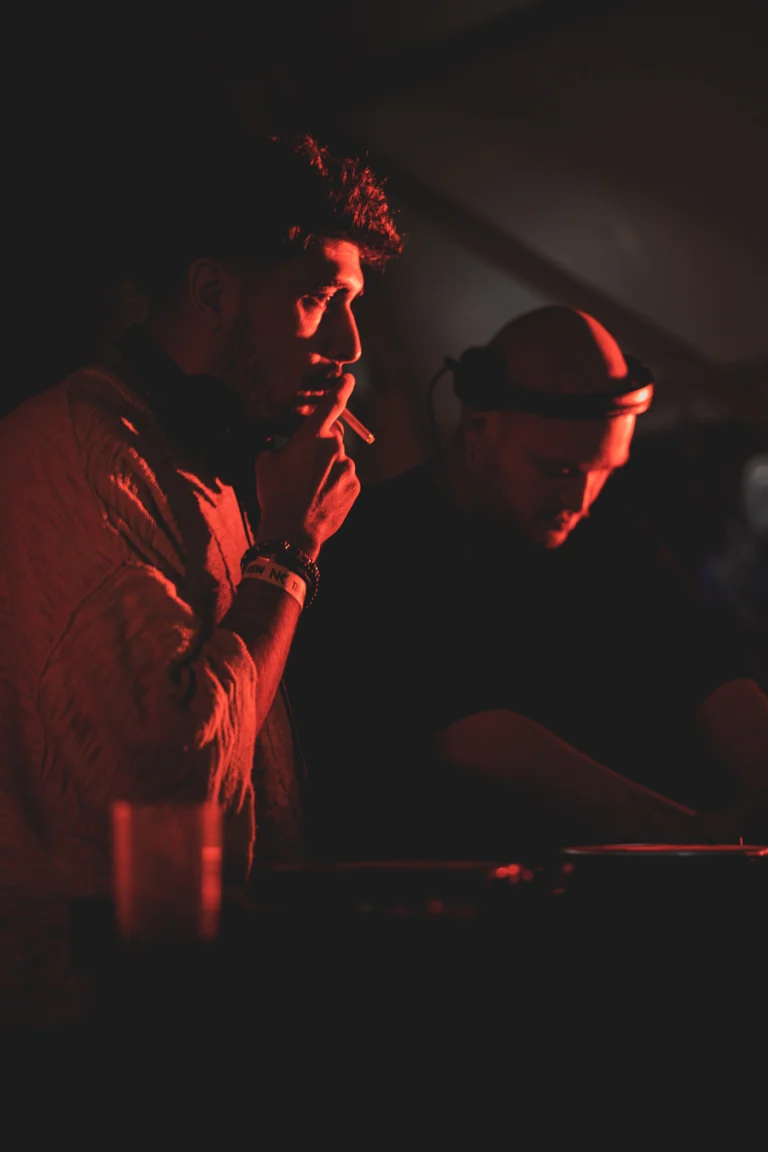 Two men performing in red club lighting, with one man holding a cigarette to his mouth and the other man standing behind a mixing console.