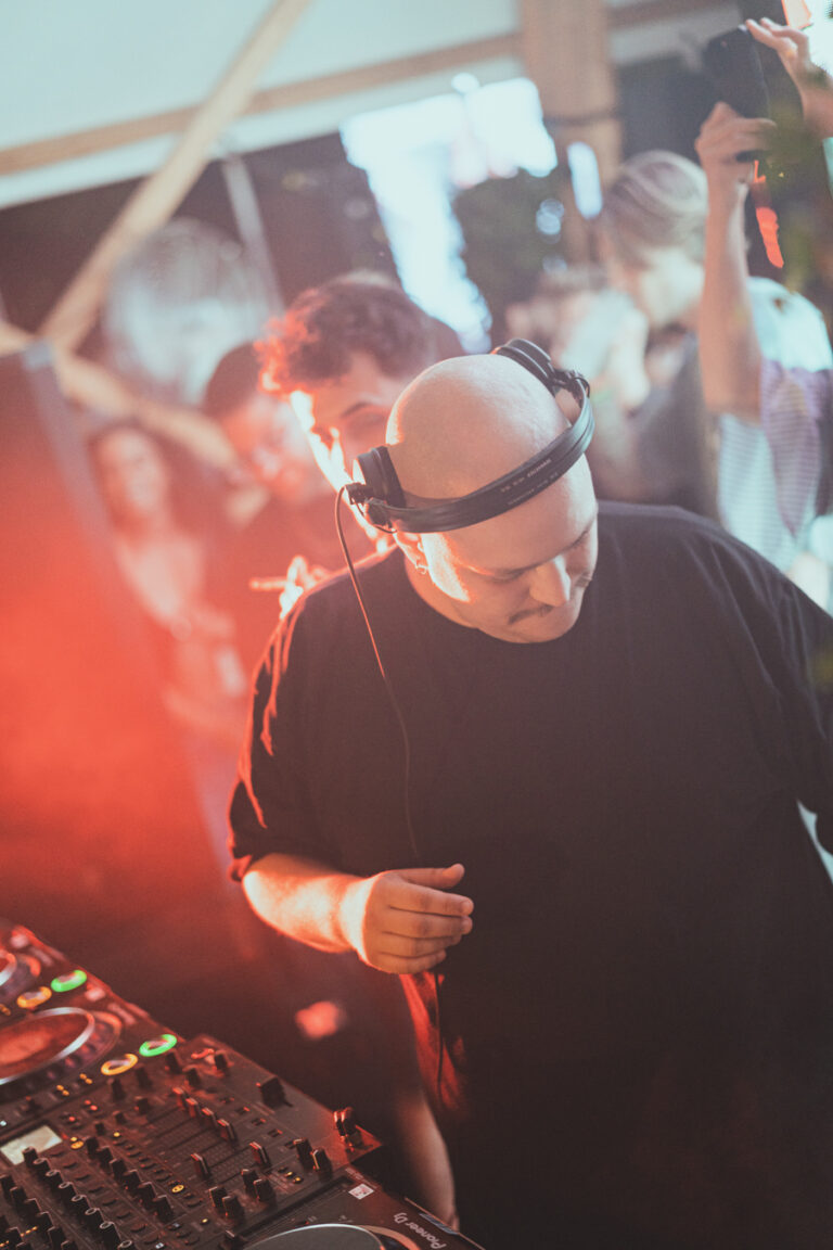 Ein kahlköpfiger Mann mit Kopfhörern um den Hals steht in warmem roten Licht hinter einem DJ-Mixer, während im Hintergrund eine Menschenmenge verschwommen zu sehen ist.