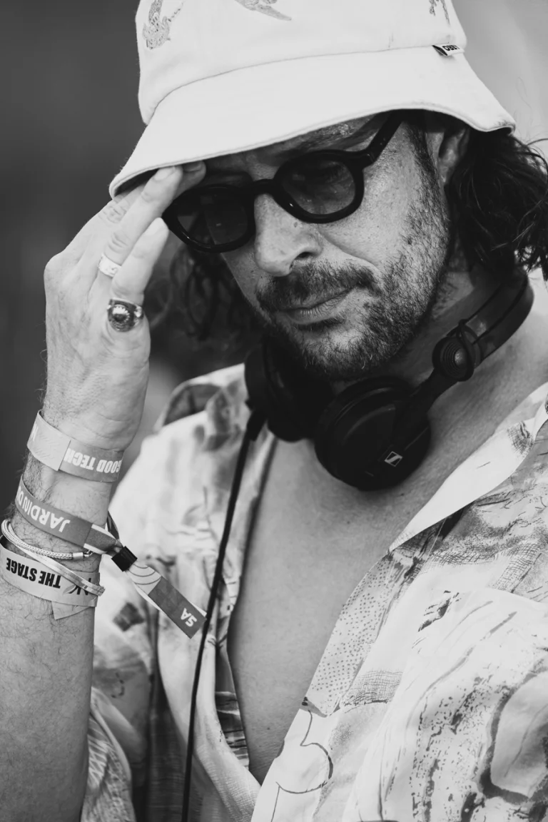 A black-and-white portrait of a man wearing a bucket hat, sunglasses, and headphones around his neck, holding his hand to the brim of his hat.