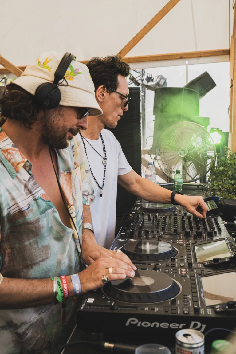 Two men operating a DJ mixer setup indoors under natural light.