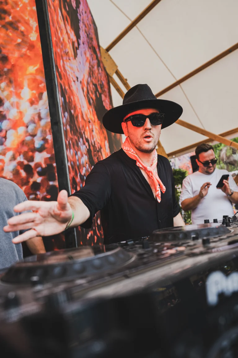 A man wearing a wide-brimmed black hat, sunglasses, and a patterned neck scarf gestures while DJing behind a mixing console.