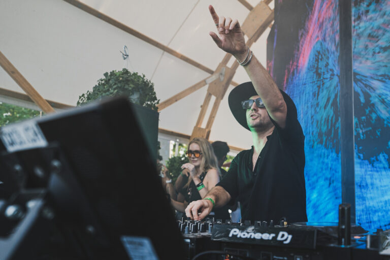 Ein Mann mit Hut und Sonnenbrille steht hinter einem Pioneer-DJ-Mixer und streckt eine Hand in die Höhe.
