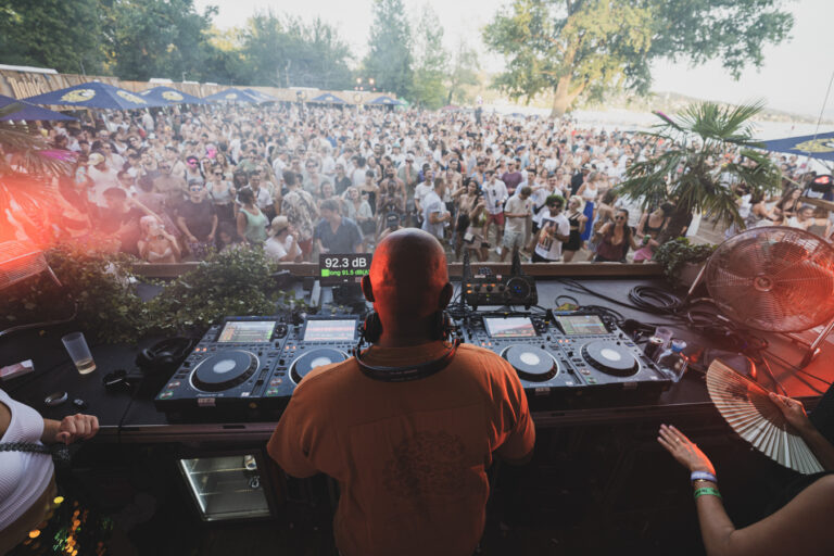 Ein kahlgänziger Mann mit dem Rücken zur Kamera steht hinter einem DJ-Pult mit Plattentepellern und blickt auf eine große Menschenmenge.