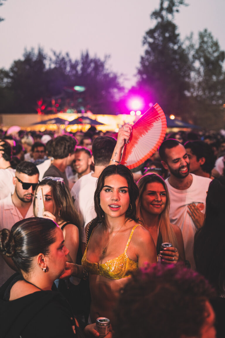 Eine Frau in einem gelben Oberteil steht in einer dichten Menschenmenge auf einem Festival im Freien unter rosa Bühnenbeleuchtung und hält einen rot-weißen Fächer hoch.