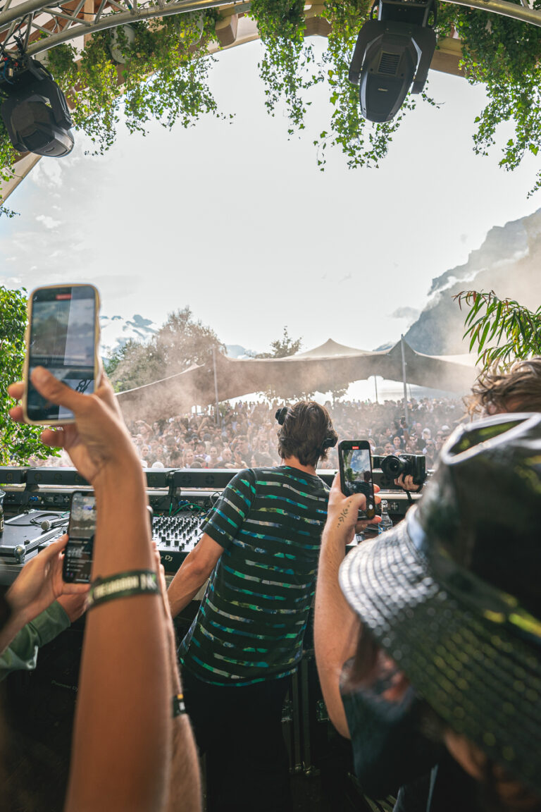 Ein Mann steht an einem DJ-Pult unter einem Blätterdach, von hinten gesehen, während Menschen in der Menge Smartphones zum Filmen hochhalten.