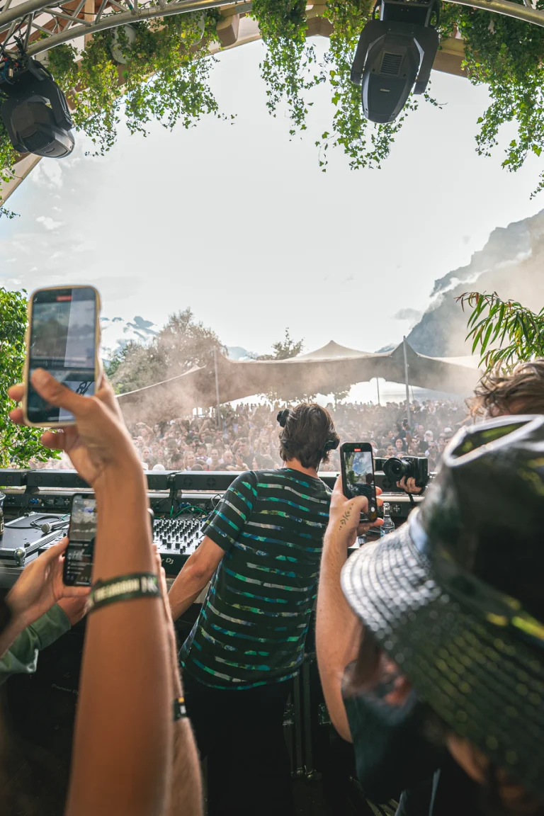 A man stands at a DJ mixer under a leafy canopy, viewed from behind as people in the crowd hold up smartphones to record.