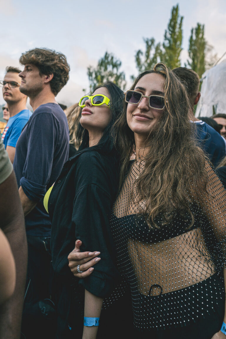 Two women stand close together in an outdoor crowd, wearing sunglasses and smiling.