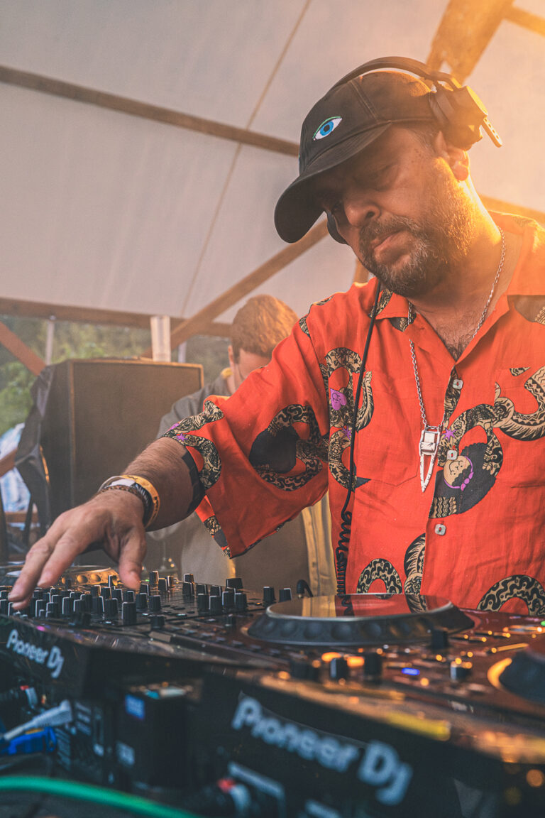 Ein Mann mit Kopfhörern und einem gemusterten orangen Hemd bedient ein Pioneer-DJ-Mischpult bei warmem Licht.