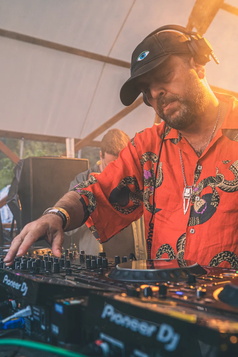 A man wearing headphones and a patterned orange shirt operates a Pioneer DJ mixing console under warm lighting.